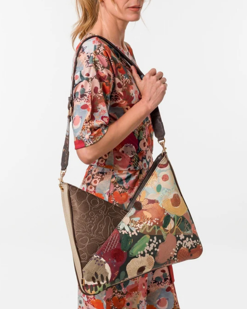 Uma carteira de ombro que celebra a cor, a arte e a liberdade numa peça de grande formato e impacto visual imediato.

O seu design em patchwork combina um painel em couro sintético castanho-escuro com bordado tonal em relevo — onde florescem motivos orgânicos e a silhueta inconfundível da personagem Anekke — com um segundo painel em impressão artística de inspiração botânica, com flores, frutos e manchas de cor em tons de laranja, verde, bege e bordô.