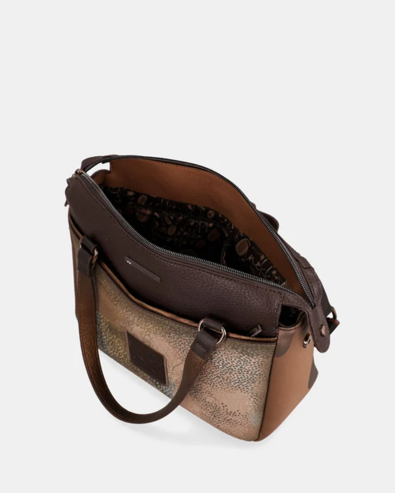Uma carteira que carrega consigo o espírito da viagem e a sofisticação do detalhe bem executado.

O seu exterior combina couro sintético em tons de bronze envelhecido e castanho-escuro, com aplicações em relevo de motivos florais gravados e silhuetas orgânicas em técnica patchwork — uma composição que evoca mapas antigos, diários de viagem e a beleza do encontro entre culturas. A assinatura Anekke em metal gunmetal e os fechos e argolas em tom escuro completam uma estética coesa e premium.