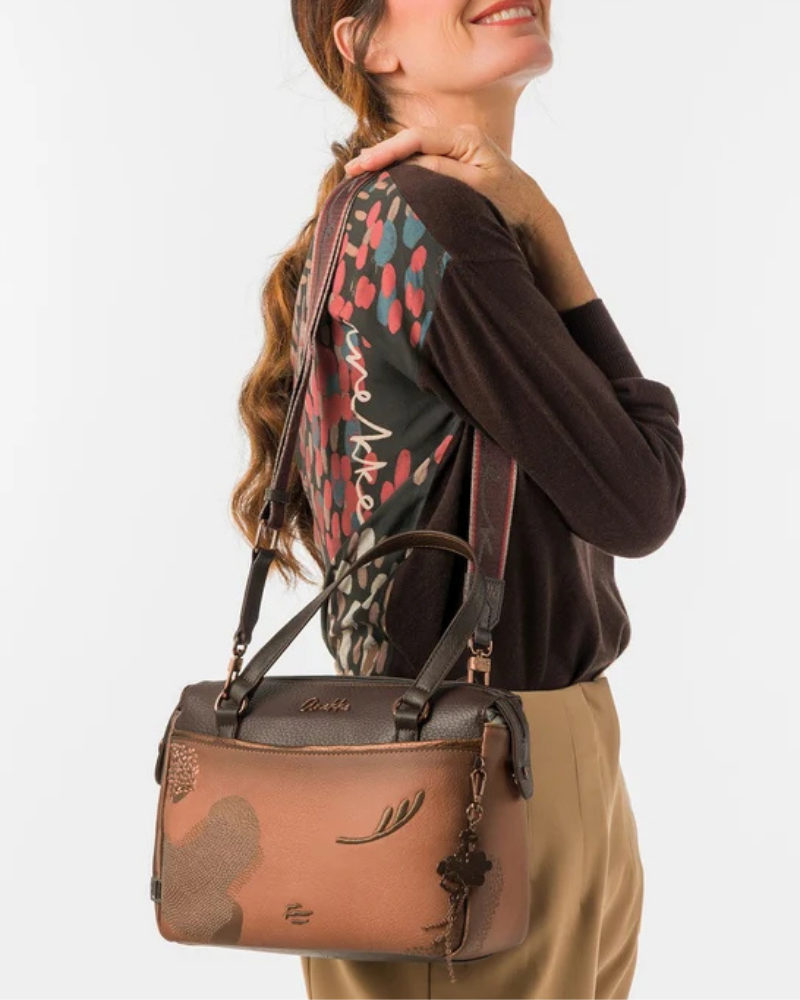 Uma carteira que carrega consigo o espírito da viagem e a sofisticação do detalhe bem executado.

O seu exterior combina couro sintético em tons de bronze envelhecido e castanho-escuro, com aplicações em relevo de motivos florais gravados e silhuetas orgânicas em técnica patchwork — uma composição que evoca mapas antigos, diários de viagem e a beleza do encontro entre culturas. A assinatura Anekke em metal gunmetal e os fechos e argolas em tom escuro completam uma estética coesa e premium.