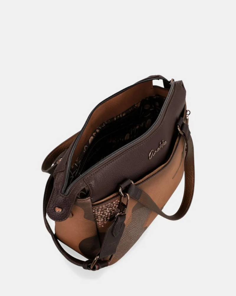 Uma carteira que carrega consigo o espírito da viagem e a sofisticação do detalhe bem executado.

O seu exterior combina couro sintético em tons de bronze envelhecido e castanho-escuro, com aplicações em relevo de motivos florais gravados e silhuetas orgânicas em técnica patchwork — uma composição que evoca mapas antigos, diários de viagem e a beleza do encontro entre culturas. A assinatura Anekke em metal gunmetal e os fechos e argolas em tom escuro completam uma estética coesa e premium.