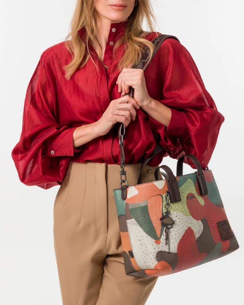 Esta carteira é muito mais do que uma carteira: é um conjunto reversível e modular que oferece duas personalidades num só produto.

A tote principal destaca-se pelo seu padrão artístico em tons de terracota, verde-musgo, laranja e creme, com formas abstratas e texturas em relevo que evocam uma pintura contemporânea. As asas duplas em castanho-escuro, os fechos e argolas em metal gunmetal e o pendente de chaves decorativo completam o design com um toque boémio e editorial.