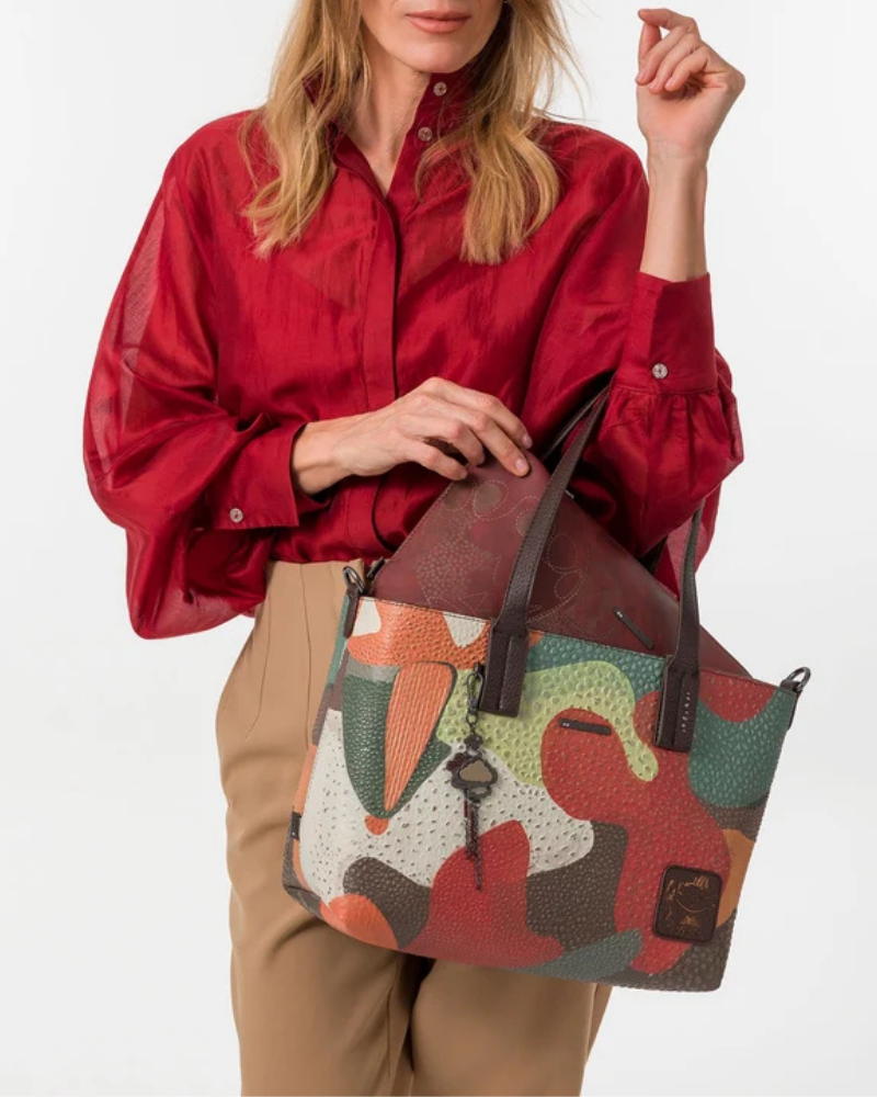 Esta carteira é muito mais do que uma carteira: é um conjunto reversível e modular que oferece duas personalidades num só produto.

A tote principal destaca-se pelo seu padrão artístico em tons de terracota, verde-musgo, laranja e creme, com formas abstratas e texturas em relevo que evocam uma pintura contemporânea. As asas duplas em castanho-escuro, os fechos e argolas em metal gunmetal e o pendente de chaves decorativo completam o design com um toque boémio e editorial.