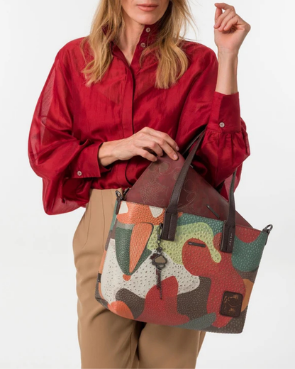 Esta carteira é muito mais do que uma carteira: é um conjunto reversível e modular que oferece duas personalidades num só produto.

A tote principal destaca-se pelo seu padrão artístico em tons de terracota, verde-musgo, laranja e creme, com formas abstratas e texturas em relevo que evocam uma pintura contemporânea. As asas duplas em castanho-escuro, os fechos e argolas em metal gunmetal e o pendente de chaves decorativo completam o design com um toque boémio e editorial.