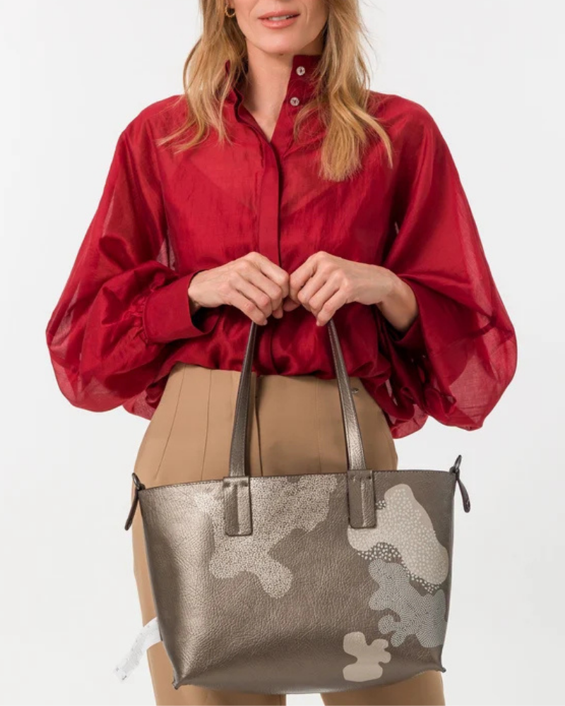 Esta carteira é muito mais do que uma carteira: é um conjunto reversível e modular que oferece duas personalidades num só produto.

A tote principal destaca-se pelo seu padrão artístico em tons de terracota, verde-musgo, laranja e creme, com formas abstratas e texturas em relevo que evocam uma pintura contemporânea. As asas duplas em castanho-escuro, os fechos e argolas em metal gunmetal e o pendente de chaves decorativo completam o design com um toque boémio e editorial.