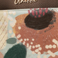 Uma celebração da natureza em forma de acessório.

O painel frontal apresenta uma ilustração botânica exuberante, com flores, folhas e formas orgânicas em tons de terracota, verde musgo, bege e coral, num estilo artístico que evoca jardins em plena floração.

A estrutura compacta e rectangular, com aba frontal e fecho de pressão dourado, é revestida em material sintético com base em verde acinzentado que ancora a composição sem competir com o estampado.